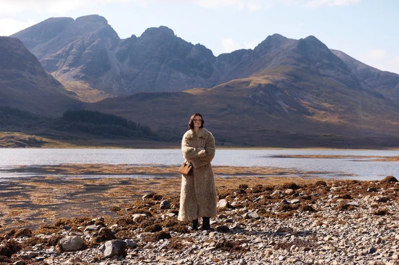 Adult woman in long coat standing on rocky shore with mountains, lake, and sky in the background on a sunny day in the outdoors.