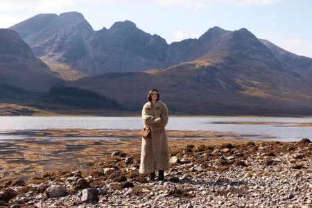 Model standing on a rocky shoreline wearing a long textured coat carrying a tan handbag against a lake and mountain backdrop