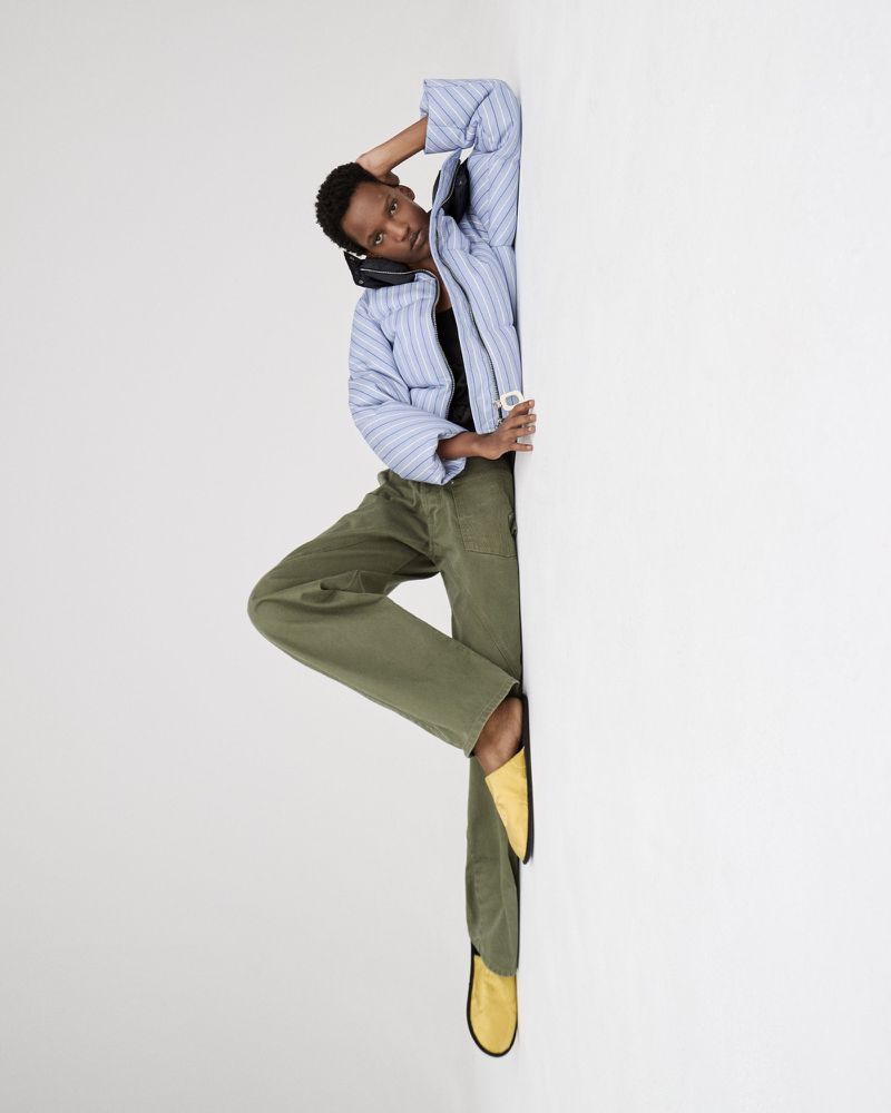 A slender, short-haired person in casual striped top and olive pants poses with headphones against a white wall, with a focus on fashion.