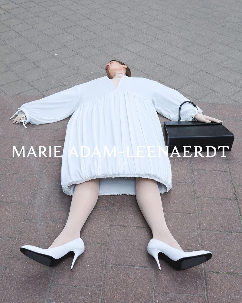 Woman in white dress and heels laying on paved ground next to a black purse, shot at eye-level with fair, diffused lighting.