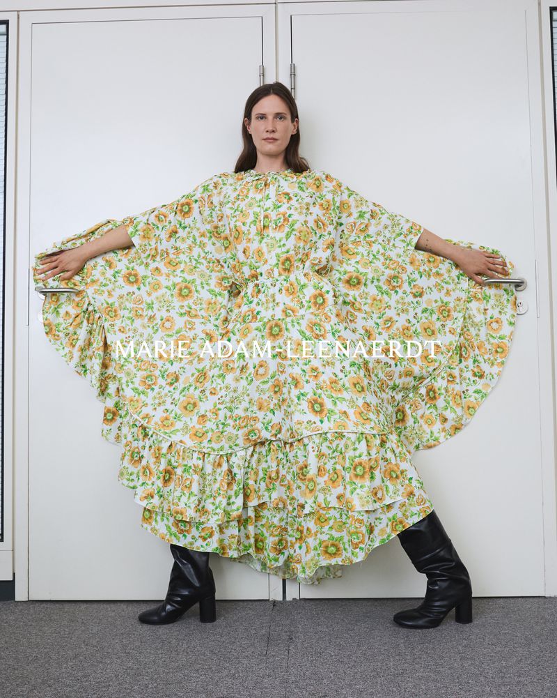 Adult female posing in a floral dress and black boots in front of white double doors with metal handles in an indoor setting.