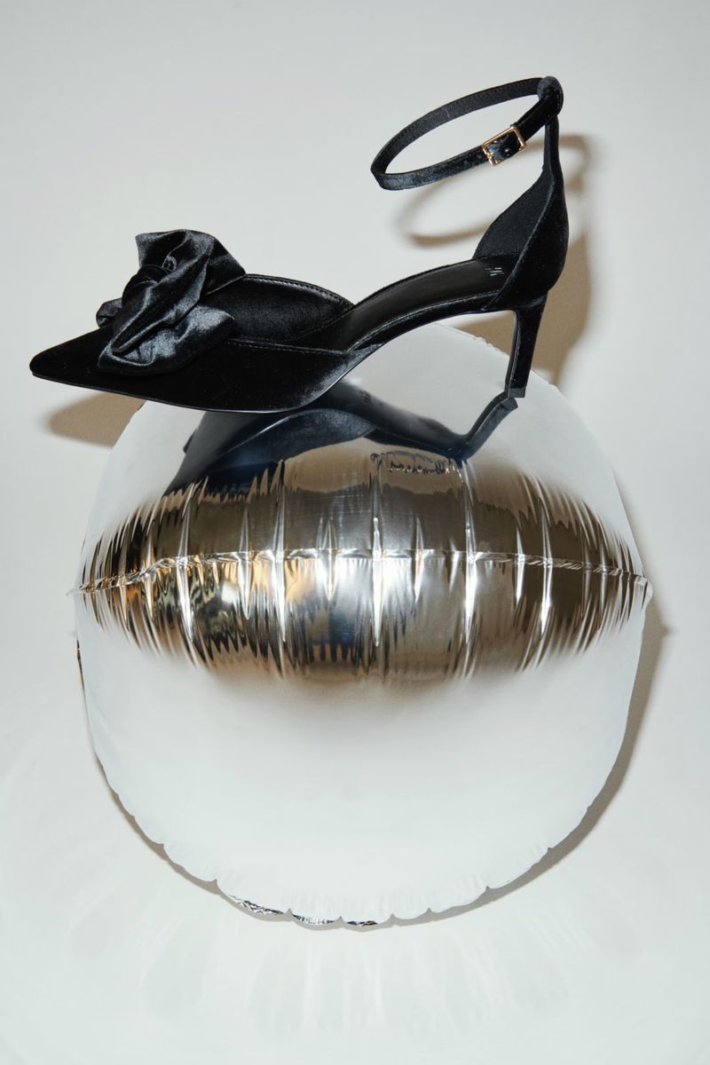 Elegant black velvet stiletto heel with bow detail perched on a silver balloon against a plain white backdrop in a fashion still life.