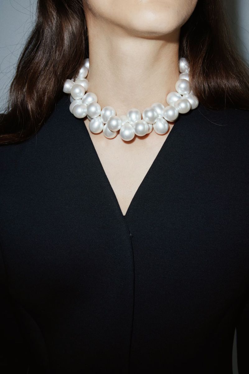 Close-up shot of a young adult woman wearing a white pearl necklace and a black V-neck top, set against a neutral background.