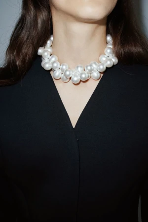 Close-up shot of a young adult woman wearing a white pearl necklace and a black V-neck top, set against a neutral background.