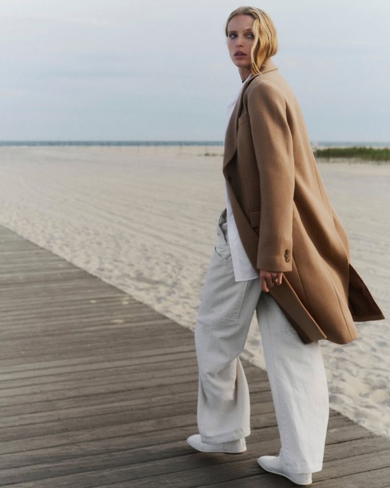 An adult female fashion model stands on a boardwalk at the beach wearing a long tan coat, wide-leg jeans, and white shoes on a cloudy day.