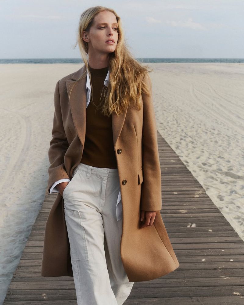 A blonde female model in a tan coat and white pants walks on a wooden boardwalk at the beach on an overcast day.
