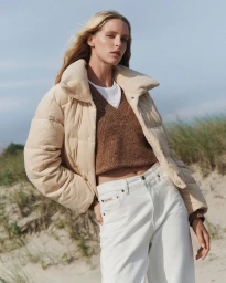 Model Abby Champion wearing a cream puffer jacket, brown sweater, and white jeans outdoors by sand dunes