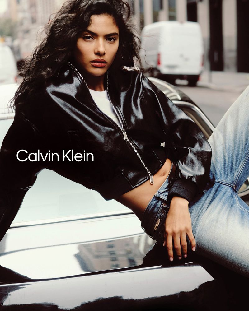 Model Rania Benchegra leaning on a car in a black leather jacket and jeans for Calvin Klein