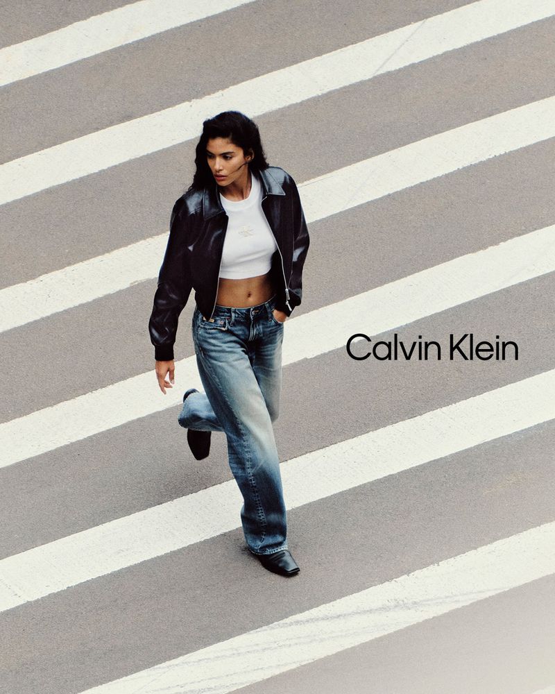 A woman in denim and a jacket walks across a crosswalk in a fashion editorial style shot for Calvin Klein, showcasing modern street style.
