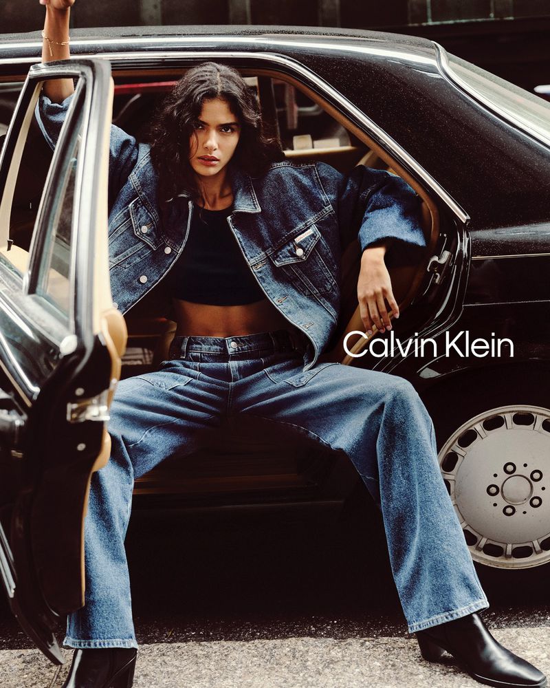 A young woman sitting inside a vintage car, wearing a denim jacket and jeans in a fashion-forward advertisement for Calvin Klein.