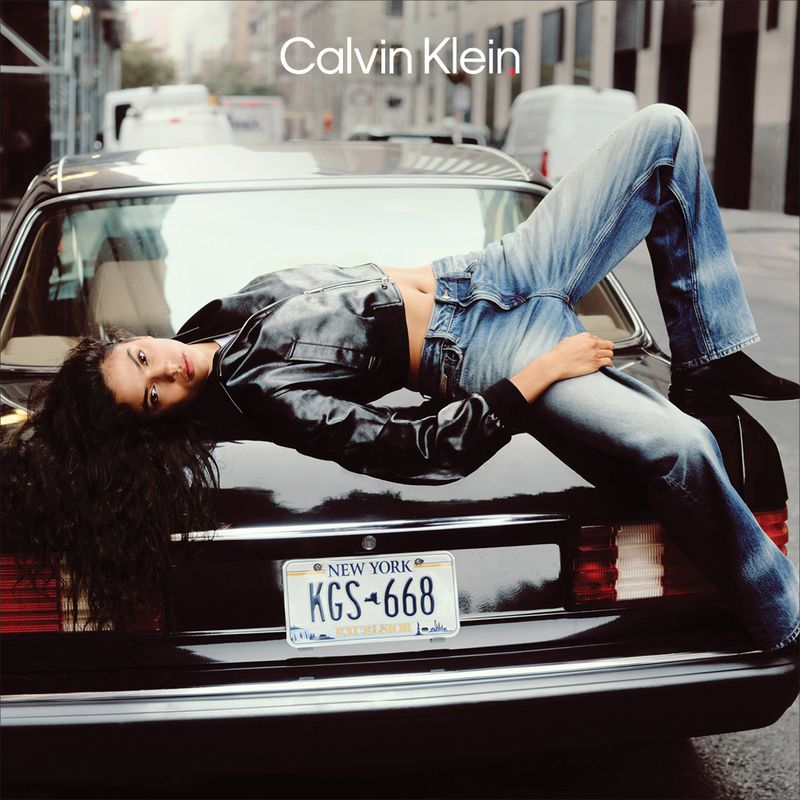 A woman lies on the trunk of a black car in a city street, wearing a black jacket and jeans, with a Calvin Klein logo visible.