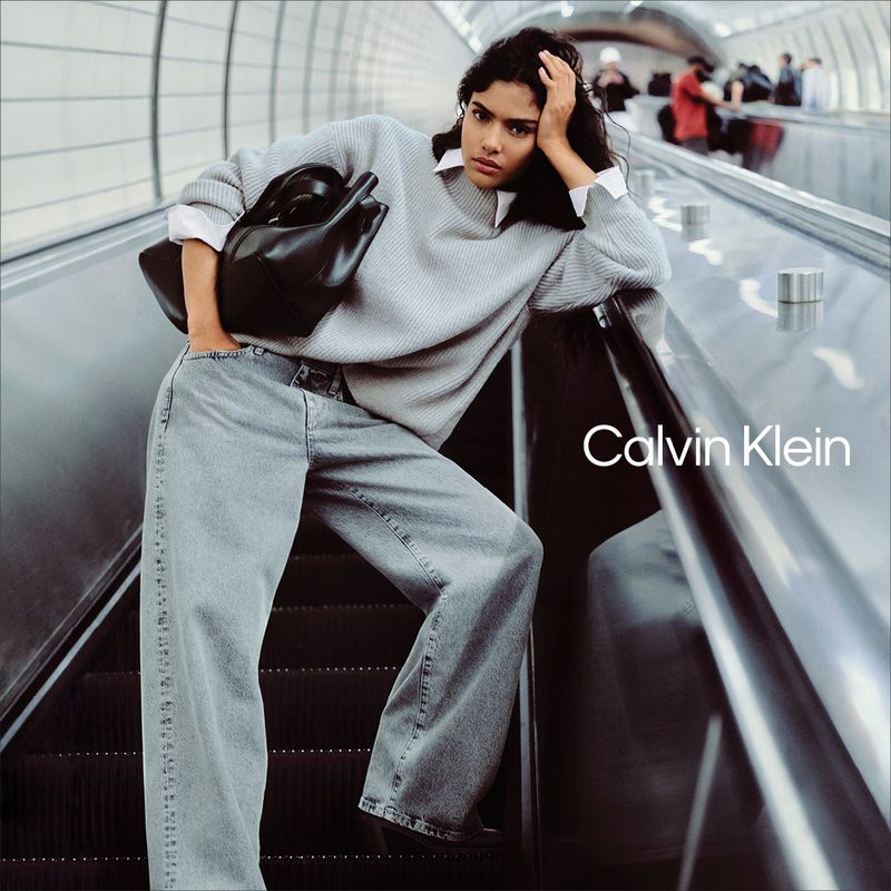 A woman poses on an escalator wearing a grey sweater, light wash denim, and a black handbag against a neutral background. 