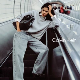 Model Rania Benchegra wearing a light grey sweater and denim jeans leaning on an escalator railing for Calvin Klein Fall 2025 Denim.