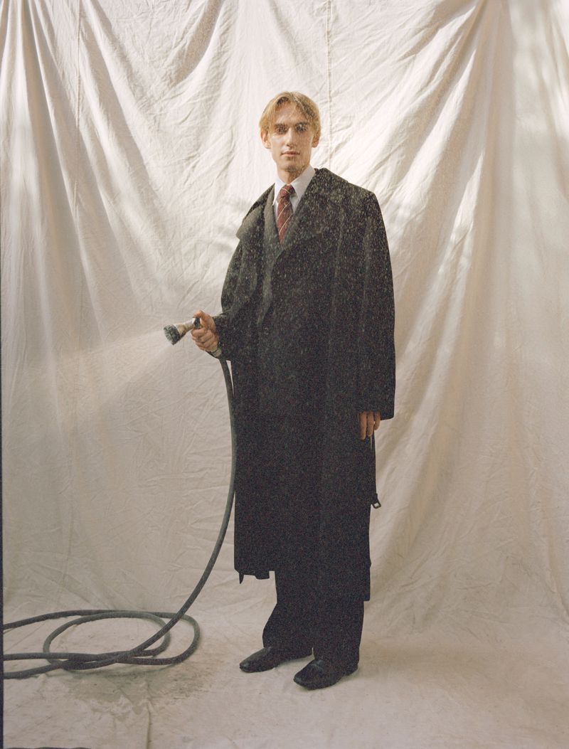 Man in overcoat holding a garden hose against a muslin backdrop in a studio setting with soft lighting. He is standing and facing forward.