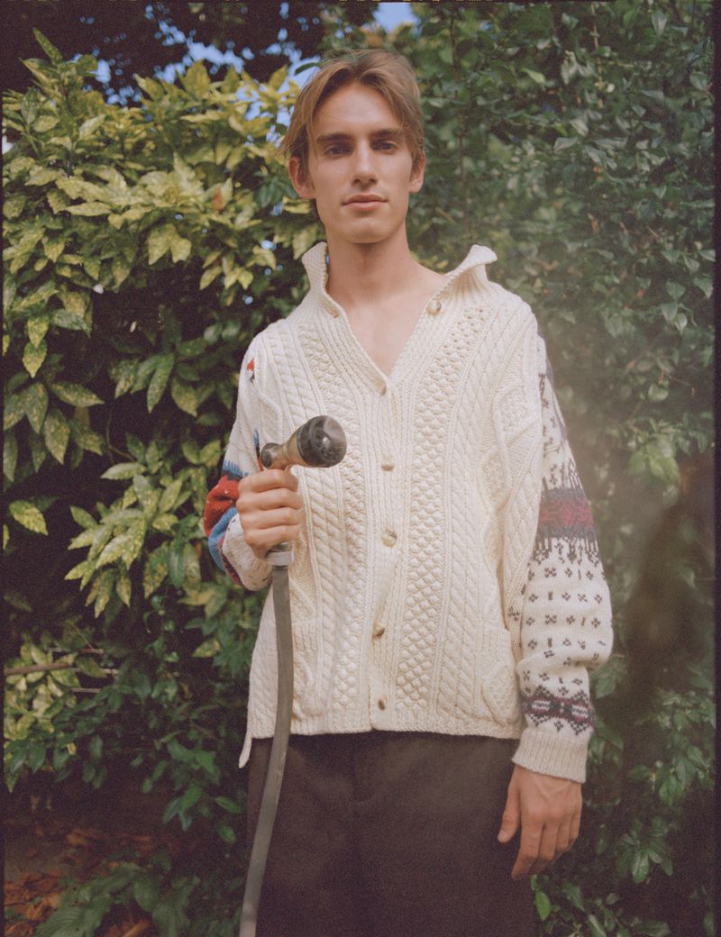 Young adult male in patterned knit sweater standing outdoors holding garden hose with lush green plants in the background.