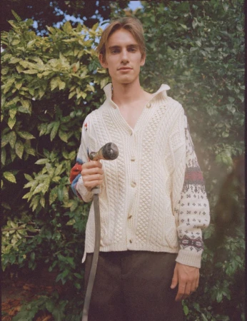 Young adult male in patterned knit sweater standing outdoors holding garden hose with lush green plants in the background.