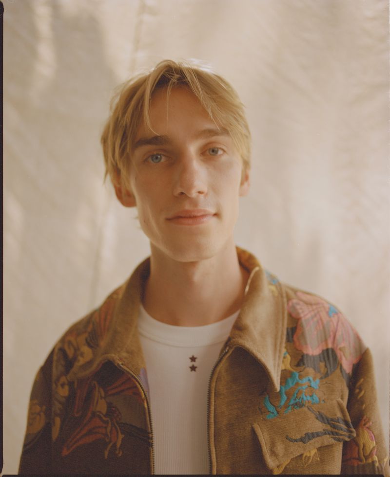 Levon Hawke wearing a patterned brown jacket and white t-shirt against a textured neutral background for Behind the Blinds