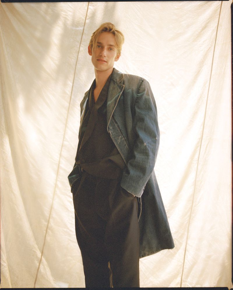 Portrait of a young adult male with blond hair wearing a long blue coat and dark pants posing in front of an off-white backdrop.