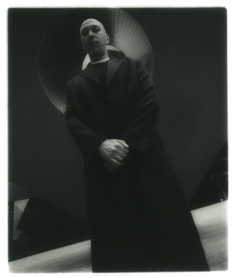 Monochromatic portrait of a bald adult male in a long black coat, looking down at the camera with geometric shapes in the background.