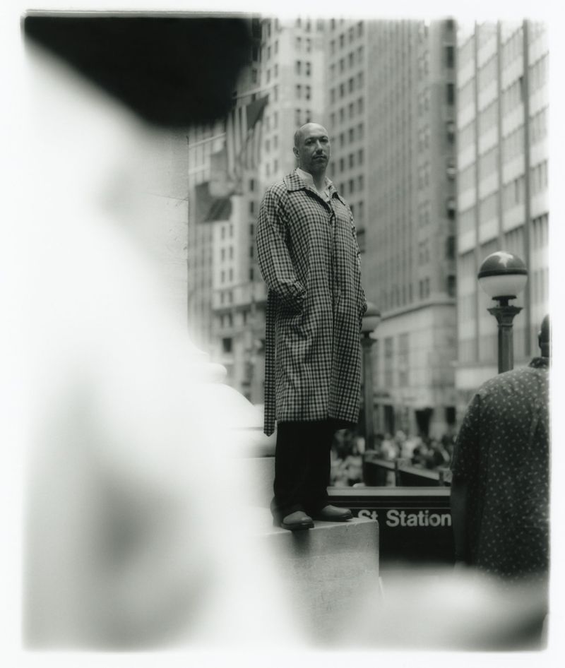 Monochrome portrait of a bald adult male in a checkered coat standing in an urban setting near a subway entrance with other people.