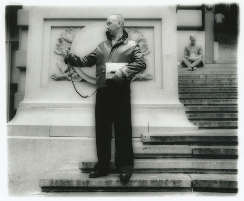 Man in jacket speaks into hand-held radio device near monument on steps. Another man sits on the steps in the background.