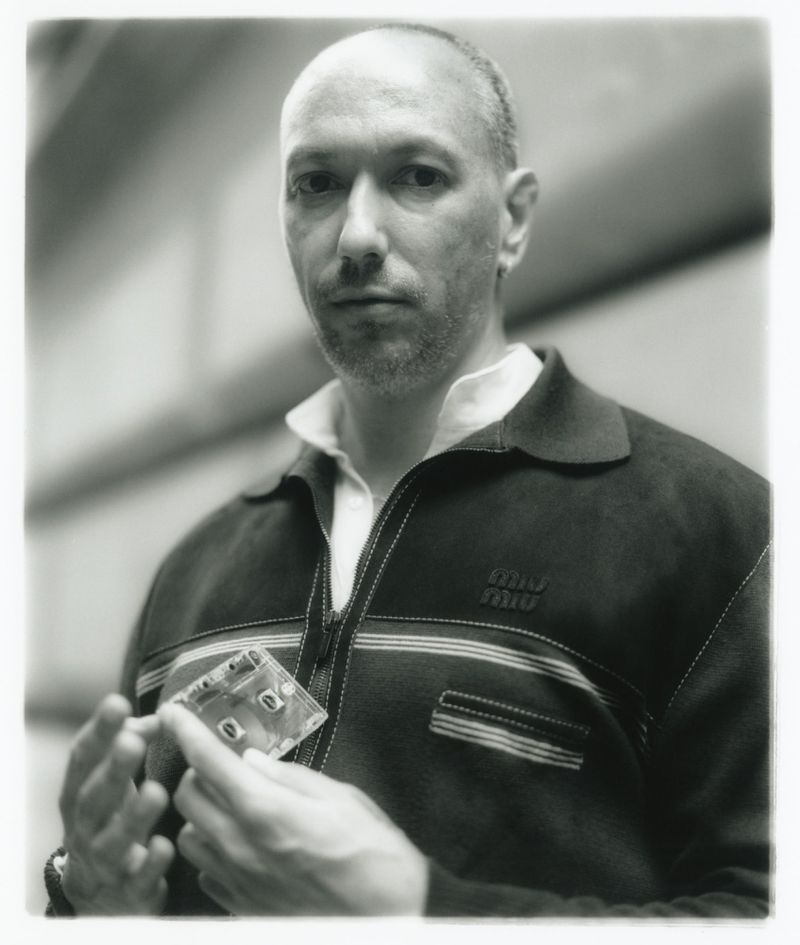 Monochrome portrait of a balding man holding a translucent cassette tape, wearing a striped shirt and jacket in an indoor environment.