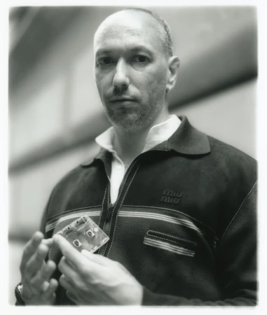Monochrome portrait of a balding man holding a translucent cassette tape, wearing a striped shirt and jacket in an indoor environment.