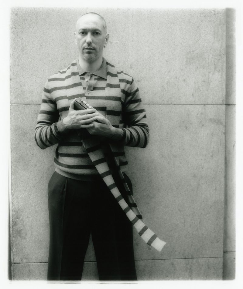 Monochrome portrait of a man in a striped shirt holding a striped scarf against a concrete wall with vignetting and soft focus.