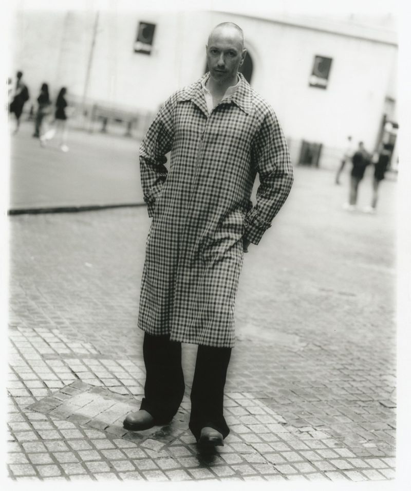 Monochrome portrait of a bald man in a patterned overcoat standing on a tiled urban plaza with blurred pedestrians in the background.