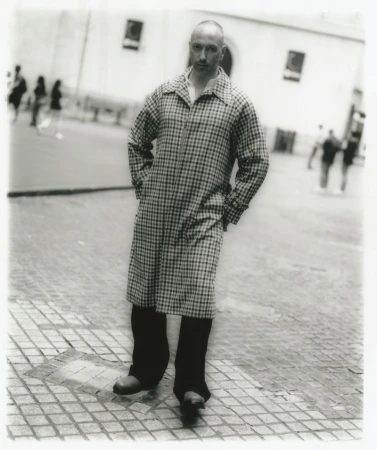 Monochrome portrait of a bald man in a patterned overcoat standing on a tiled urban plaza with blurred pedestrians in the background.