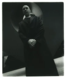 Portrait of a bald man wearing a dark coat with hands clasped, photographed in a moody interior setting by Javier Castán.