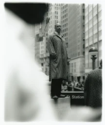 A bald man wearing a plaid patterned coat standing outdoors against a city street backdrop with a soft out-of-focus foreground blur.