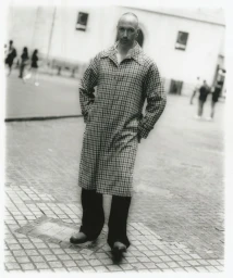 Monochrome portrait of a man standing outdoors in a plaid coat with hands in pockets on a cobbled square.