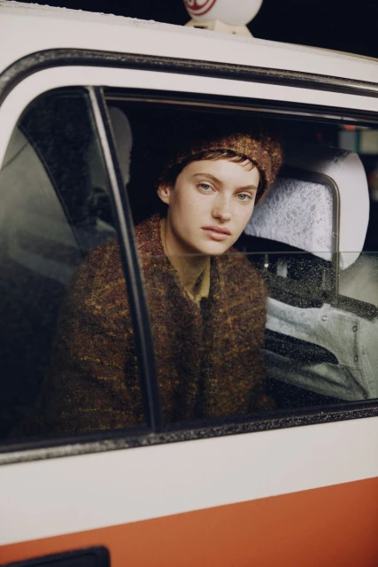 Portrait of a young adult woman with short hair and brown beret, inside a taxi with a window and part of the car visible.