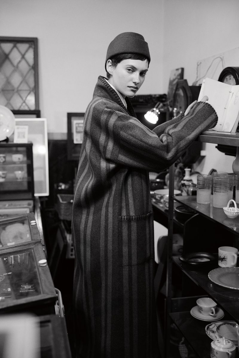 Black and white shot of a stylish adult woman wearing a striped coat and hat, browsing items on a glass shelf in a vintage store.
