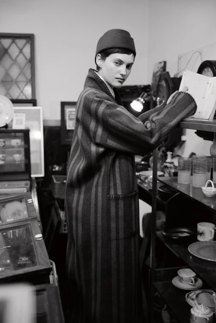 Black and white shot of a stylish adult woman wearing a striped coat and hat, browsing items on a glass shelf in a vintage store.