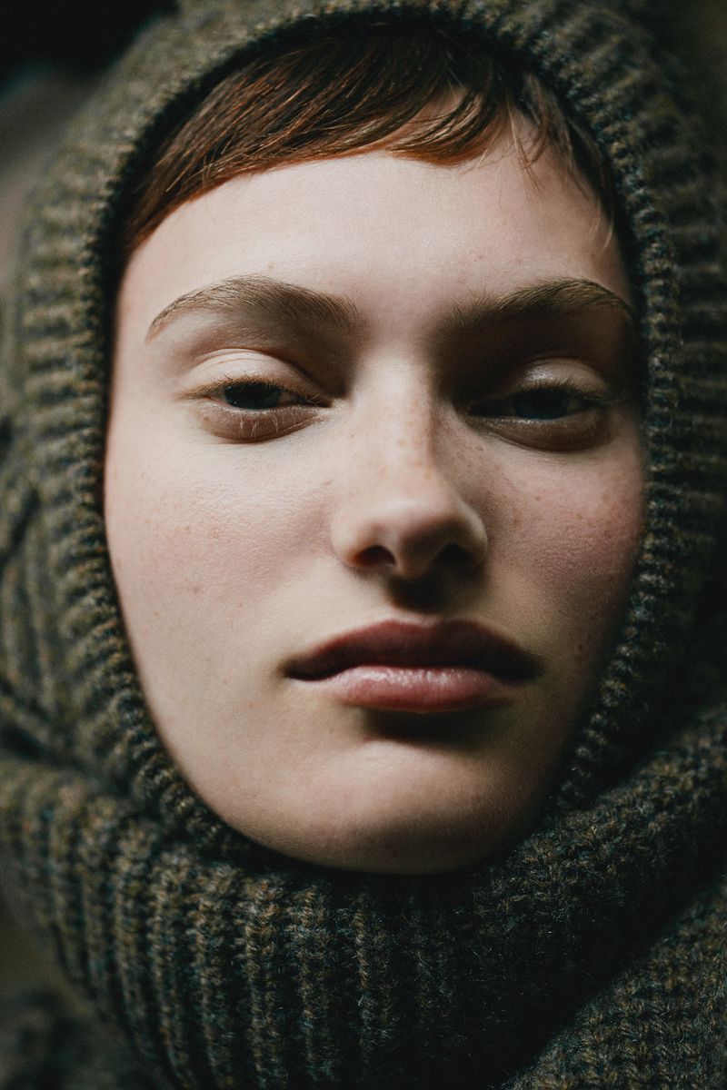 Close-up portrait of a young woman with a neutral expression wearing a textured, hooded knit hat in muted, natural light.
