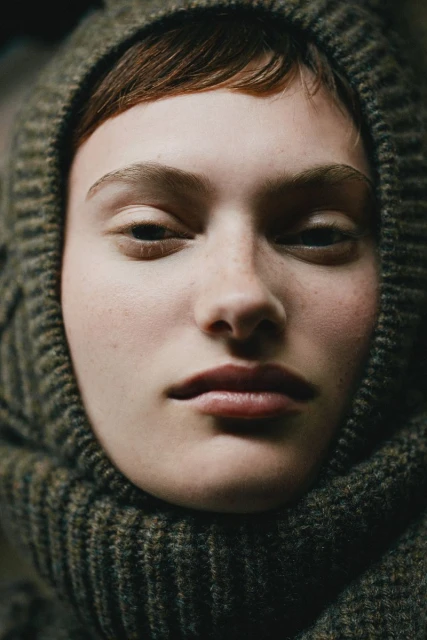 Close-up portrait of a young woman with a neutral expression wearing a textured, hooded knit hat in muted, natural light.
