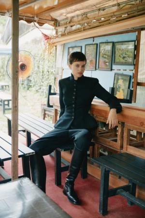 Model with short dark hair in a black buttoned jacket and tall leather boots seated at an outdoor wooden table.