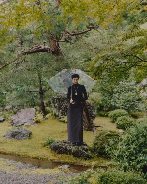 Woman in elegant black dress poses with transparent umbrella in a serene garden setting with lush green foliage and rocks.