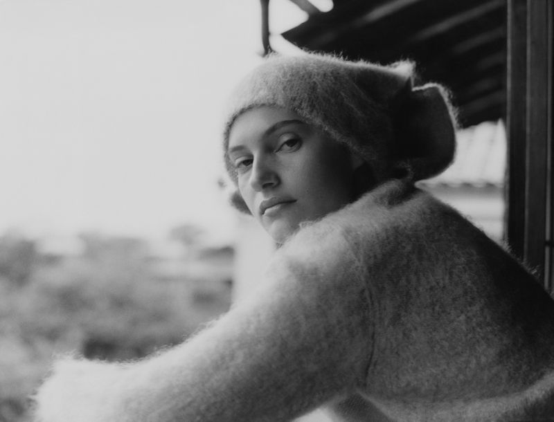 Monochrome portrait of a woman wearing a fuzzy hat and sweater looking at the camera, standing near a window with a blurred background.