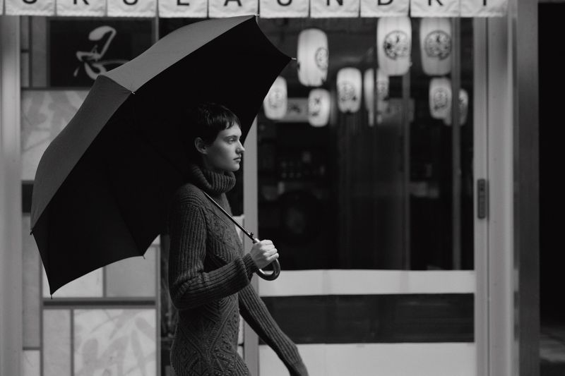 Monochrome shot of a stylish adult woman with short hair holding an open umbrella in front of a building with lanterns.