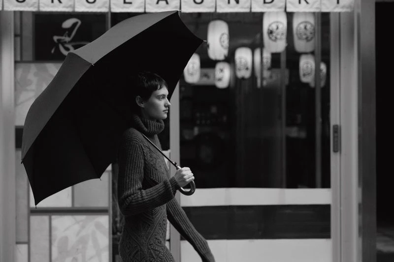 Monochrome shot of a stylish adult woman with short hair holding an open umbrella in front of a building with lanterns.