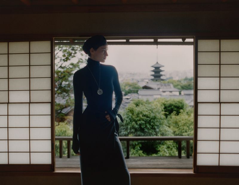An adult female stands indoors looking out an open shoji window in Japan, wearing a black dress, with trees and a pagoda in the background.