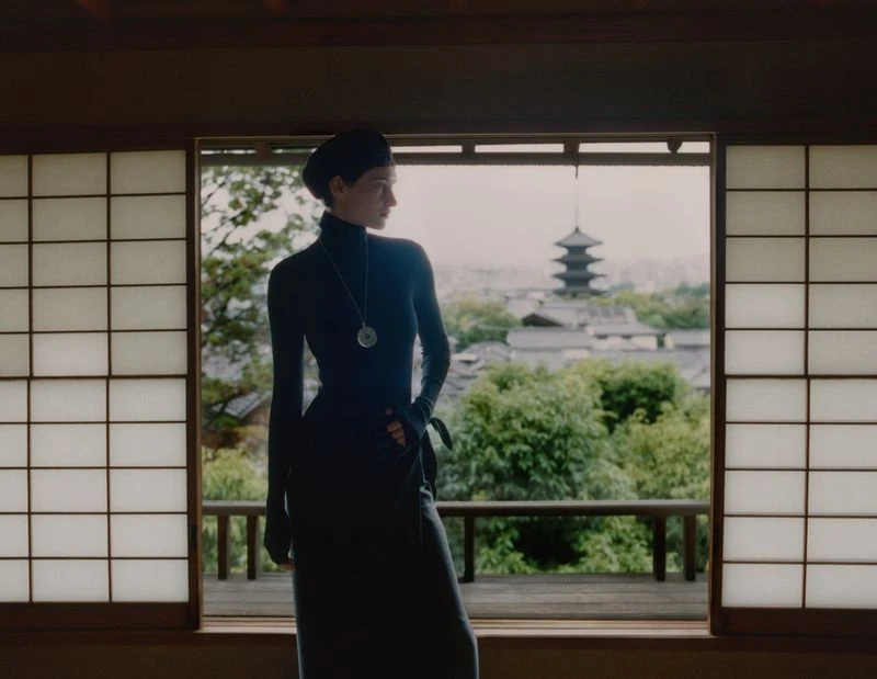 An adult female stands indoors looking out an open shoji window in Japan, wearing a black dress, with trees and a pagoda in the background.