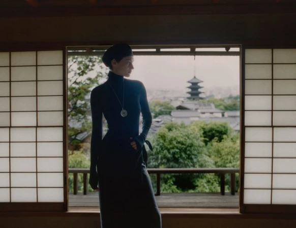 An adult female stands indoors looking out an open shoji window in Japan, wearing a black dress, with trees and a pagoda in the background.