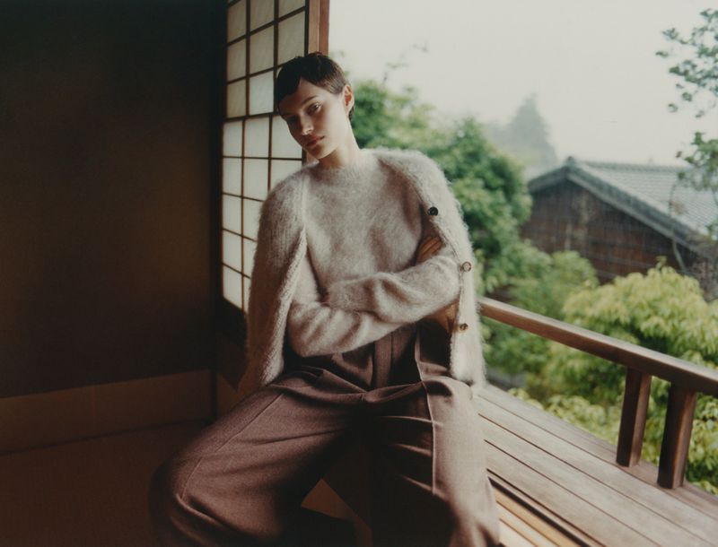 A woman with short hair poses inside with natural light, wearing a fuzzy sweater and wide-leg pants, gazing toward the camera.