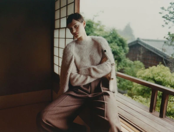 A woman with short hair poses inside with natural light, wearing a fuzzy sweater and wide-leg pants, gazing toward the camera.