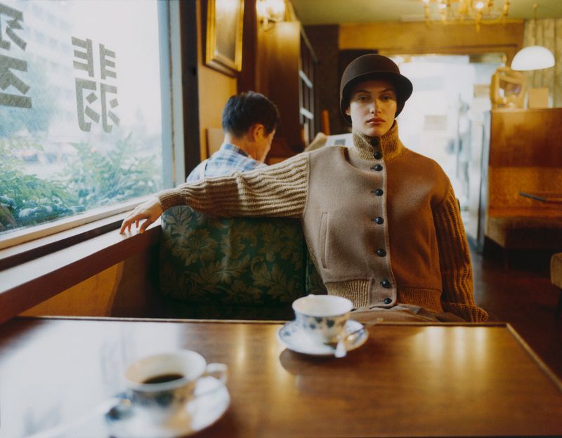 Woman wearing a brown jacket and hat sits at a cafe table with a cup of coffee, with a man in the background by a window.