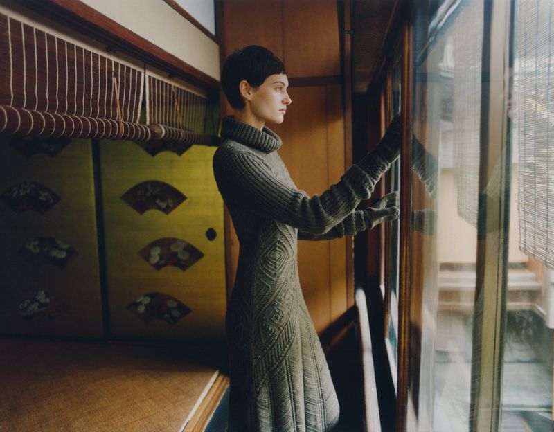 Woman in a gray dress gazes out a window in a traditionally styled Japanese interior with wooden walls and a decorative gold screen.